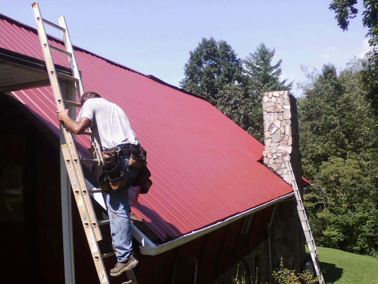 Roofer on a ladder beside a red metal roof and stone chimney — Soffit & Fascia Repair by Roofing Brothers in Wellersburg